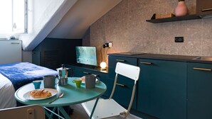 A compact kitchen with teal cabinetry, a small dining table, and a chair. There is a window providing natural light, and a plant on a shelf above the counter. - Torino Centro, Il Principino (Torino)