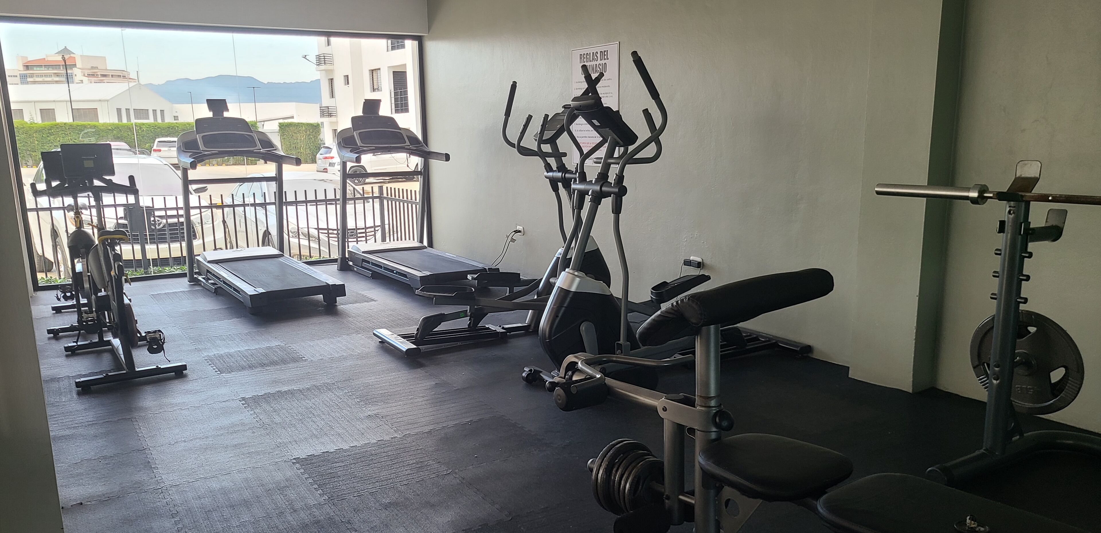 Un gimnasio con máquinas de cardio, equipo de levantamiento de pesas y un pequeño espacio exterior con vistas a edificios y montañas.