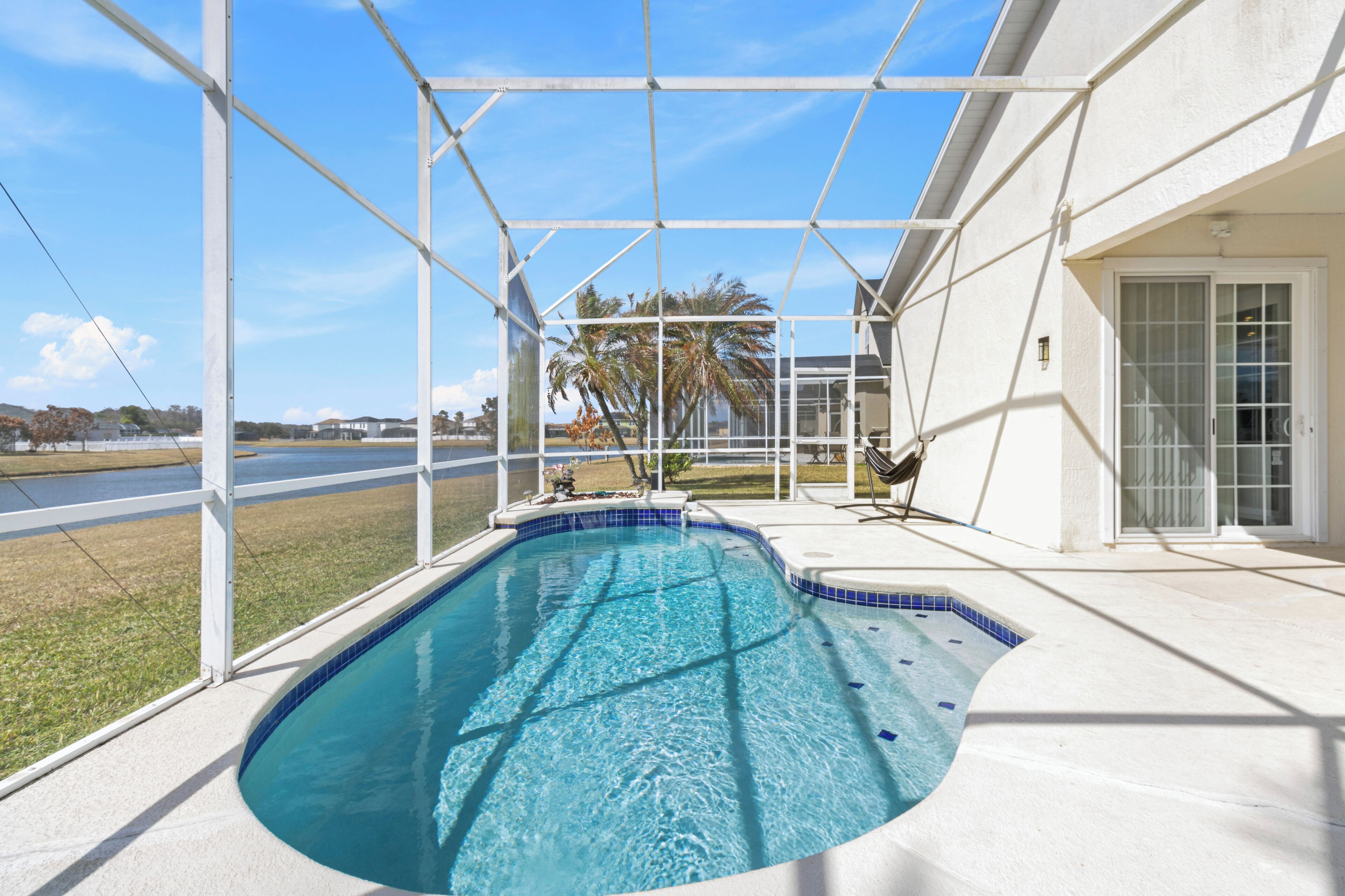A swimming pool with a clear blue water, surrounded by a white fence, and a view of palm trees and a road in the background.