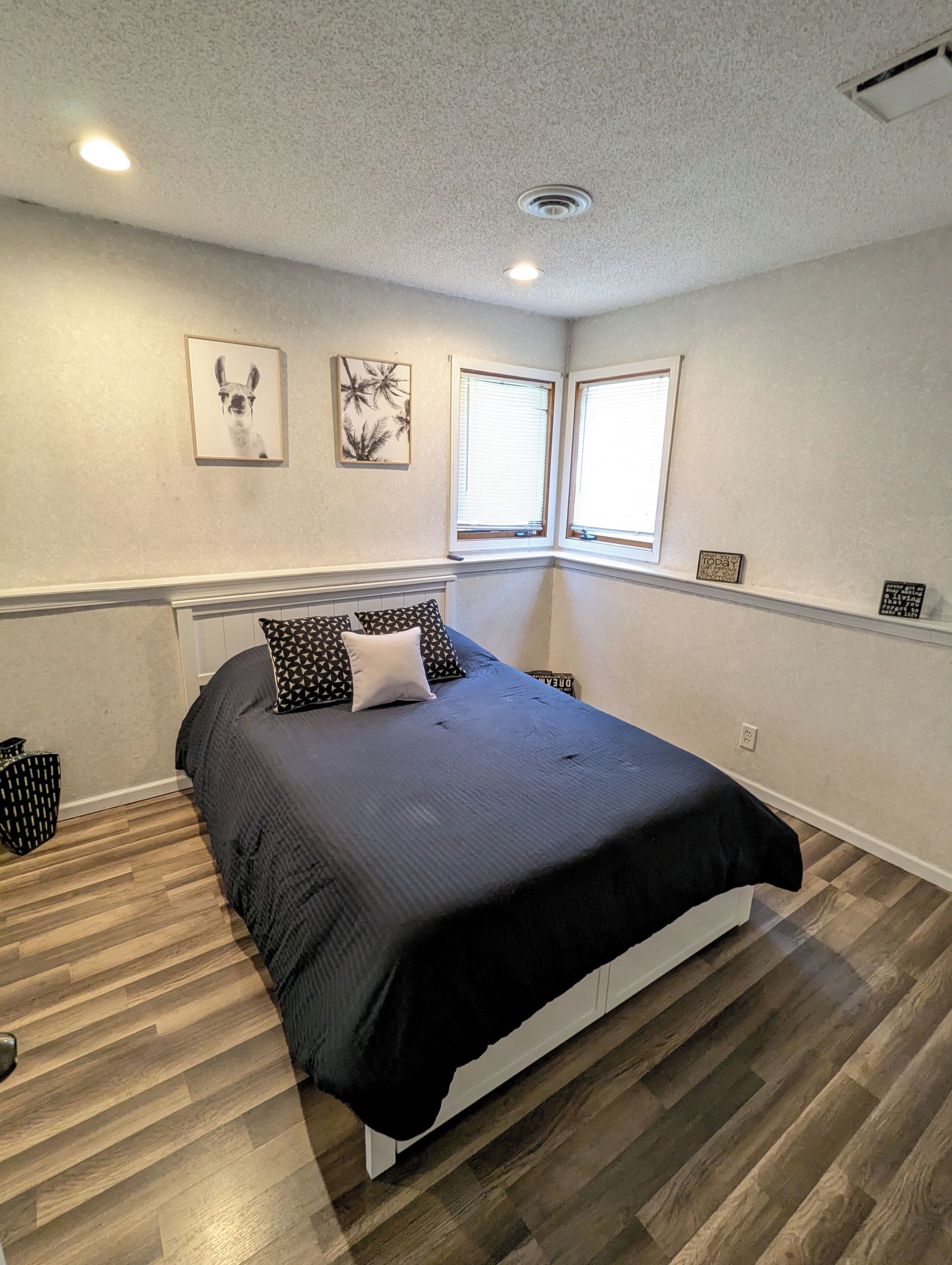 Une chambre à coucher avec un lit, deux fenêtres, un luminaire au plafond et un mur orné de tableaux encadrés.