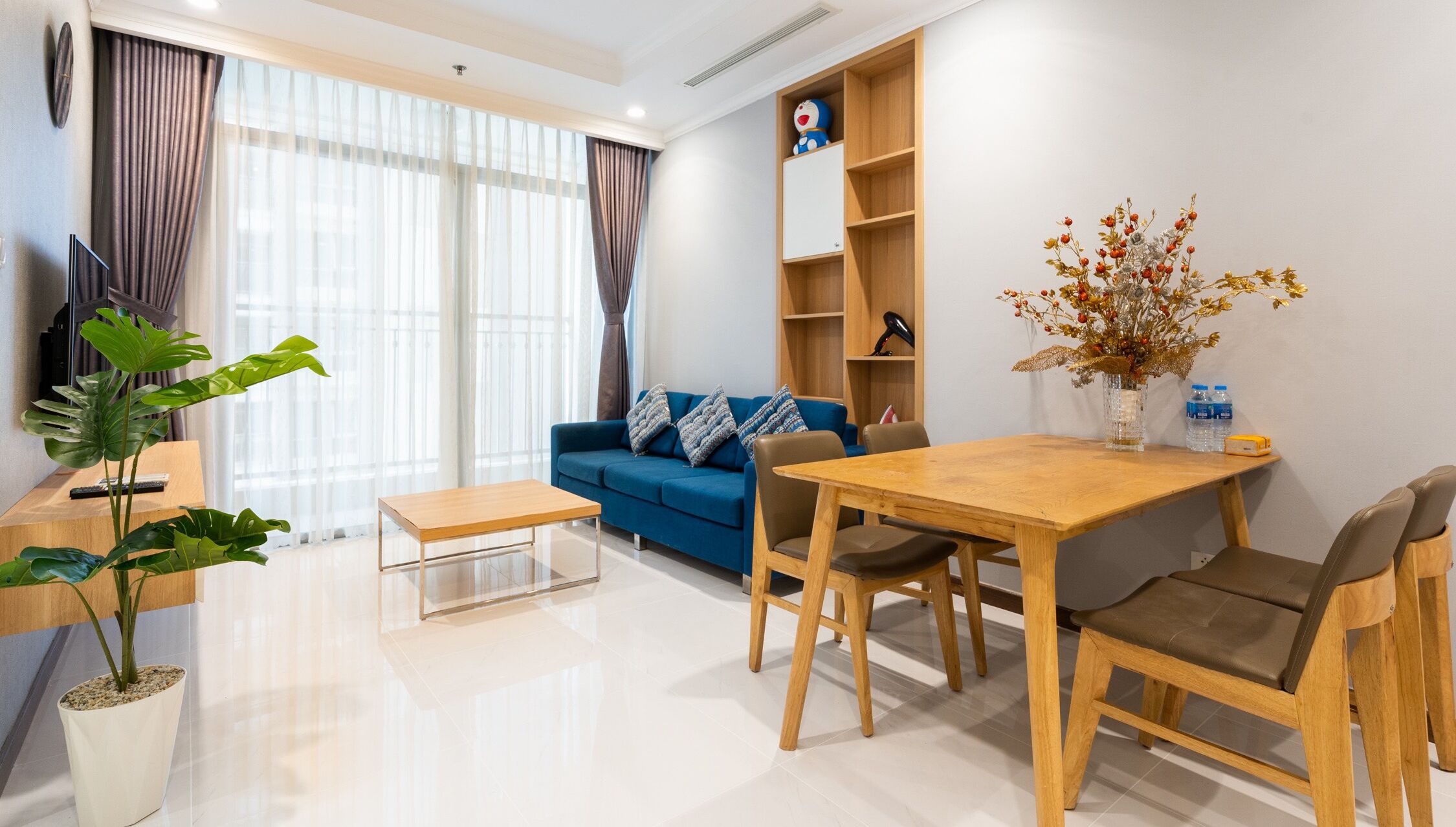 A modern living room with a blue sofa, wooden coffee table, and chairs. There is a plant, a bookshelf, and a vase with flowers.