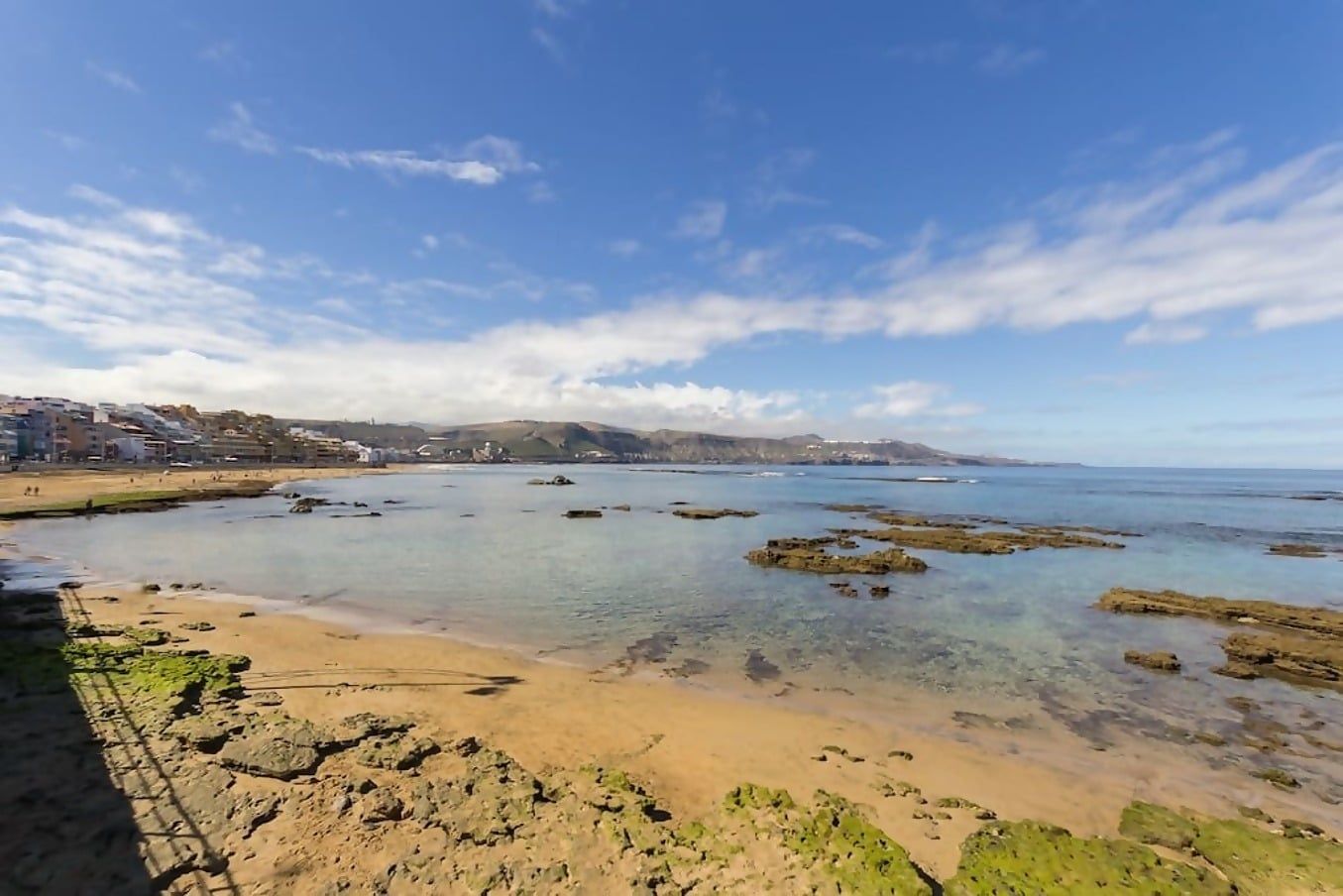 A coastal landscape with clear water, rocky formations, and buildings in the background.