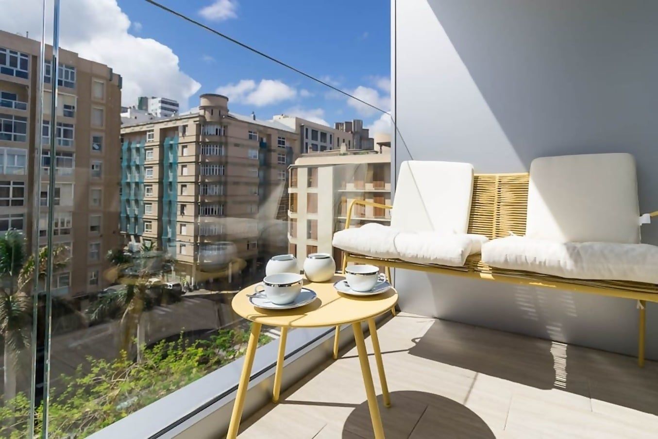 A balcony with a table set for two, a wicker chair, and a view of surrounding buildings.