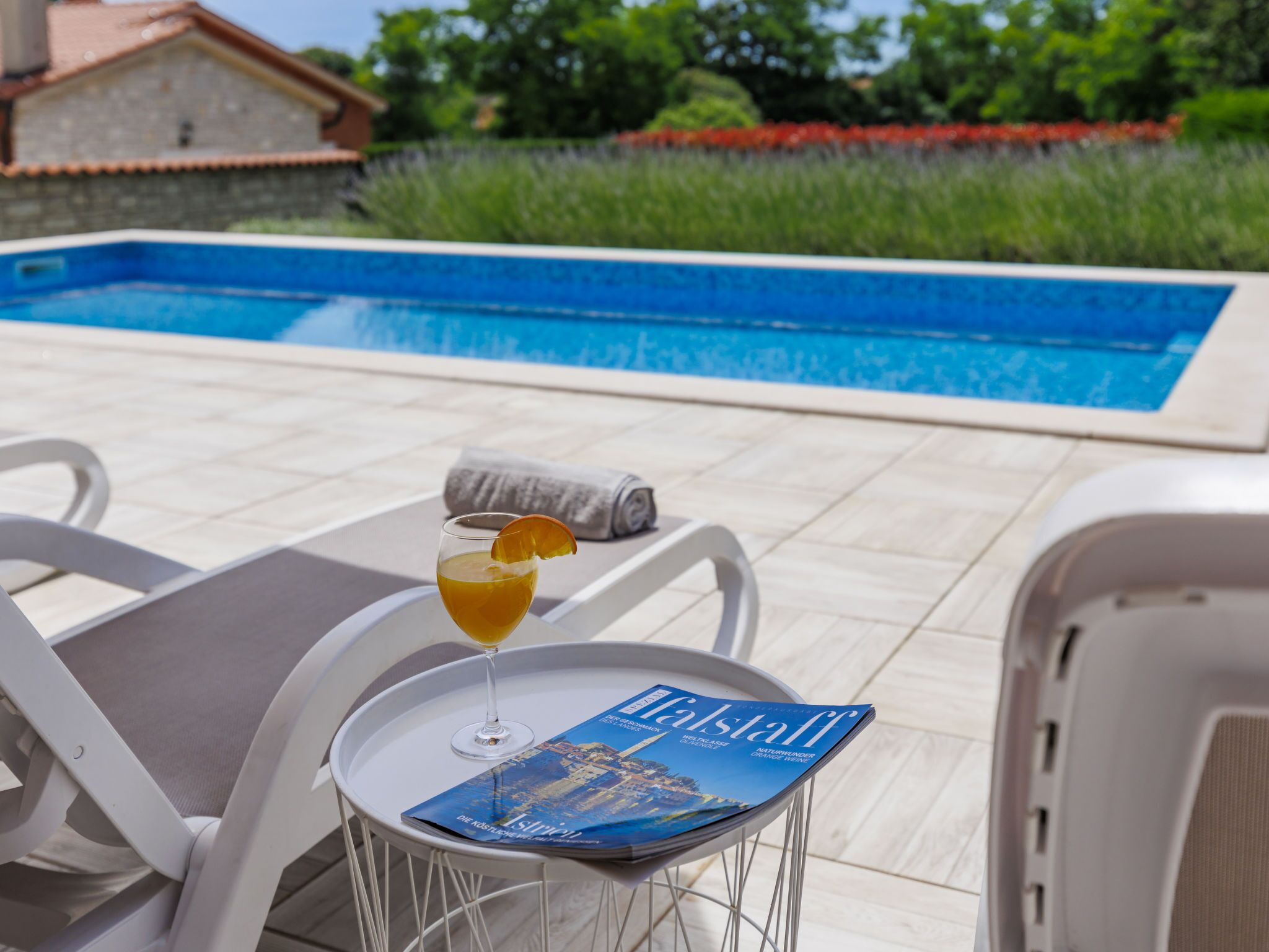 A poolside area with a lounge chair, a table with a magazine, and a glass of drink.