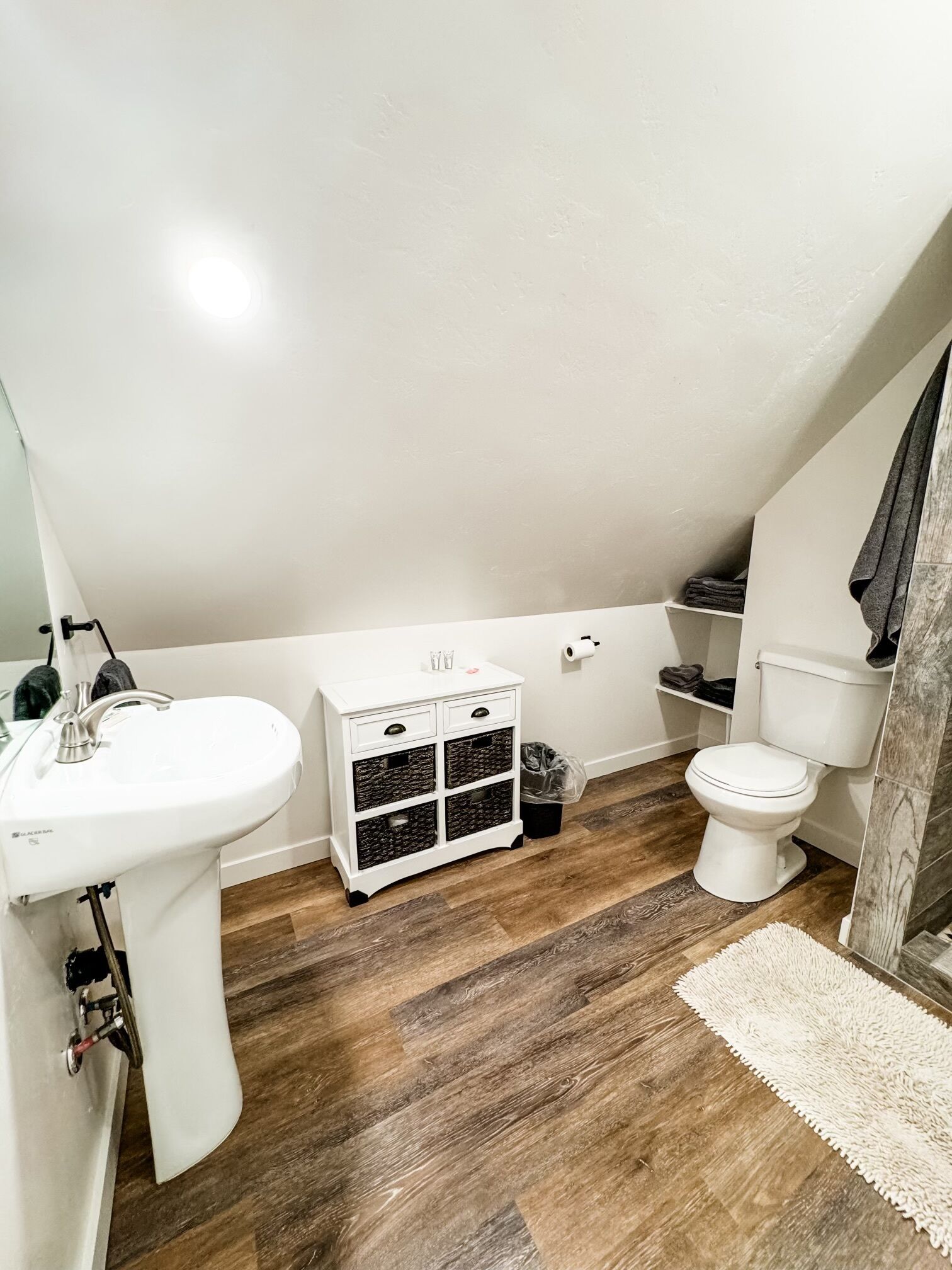 Une salle de bain avec un lavabo blanc, des toilettes, un petit meuble blanc avec des tiroirs et un plancher en bois.
