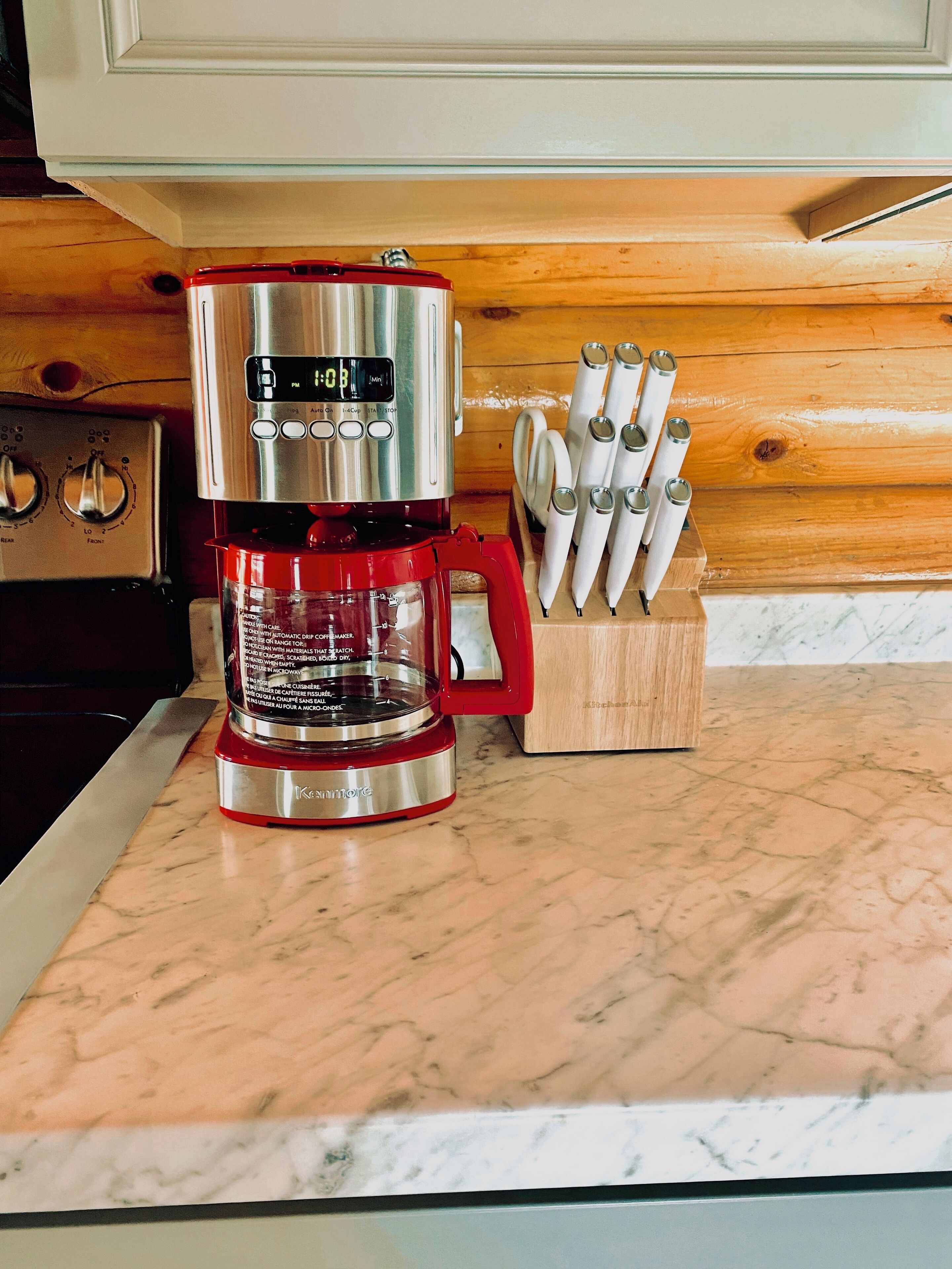 Une cuisine avec une machine à café Keurig rouge posée sur un comptoir en marbre, une planche à découper en bois avec des ustensiles, et un meuble blanc en arrière-plan.