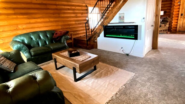 L'intérieur d'une cabane en rondins, avec un canapé en cuir vert, une table basse en bois et un escalier menant à un étage supérieur.