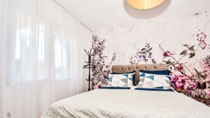 Een slaapkamer met een bed, een raam met dunne gordijnen, een bloemen behang en een nachtkastje met een lamp.