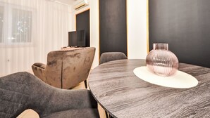 Een moderne eetruimte met een ronde tafel, grijs beklede stoelen, een decoratieve vaas en een televisietoestel op een standaard.