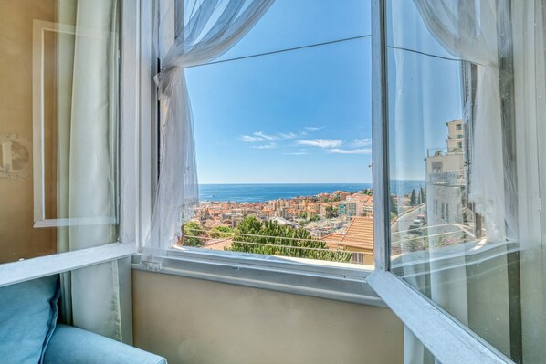Ein Zimmer mit Blick auf eine Küstenstadt durch ein Fenster mit transparenten Vorhängen.