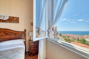 Ein Schlafzimmer mit einem Holzkopfteil, einem Bett mit weißer Bettwäsche, einem Nachttisch, einem Fenster mit durchsichtigen Vorhängen und Blick auf die Stadt.