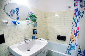 Ein Badezimmer mit Waschbecken, Spiegel, blauem Seifenspender, Zimmerpflanze und Badewanne.