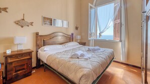 Een net opgemaakt bed met een gewatteerde deken, een houten nachtkastje met een lamp, een vis decoratie aan de muur en een raam met gordijnen.