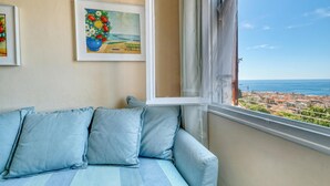 Een kamer met uitzicht op zee, een bank met blauwe kussens, en twee ingelijste kunstwerken aan de muur.