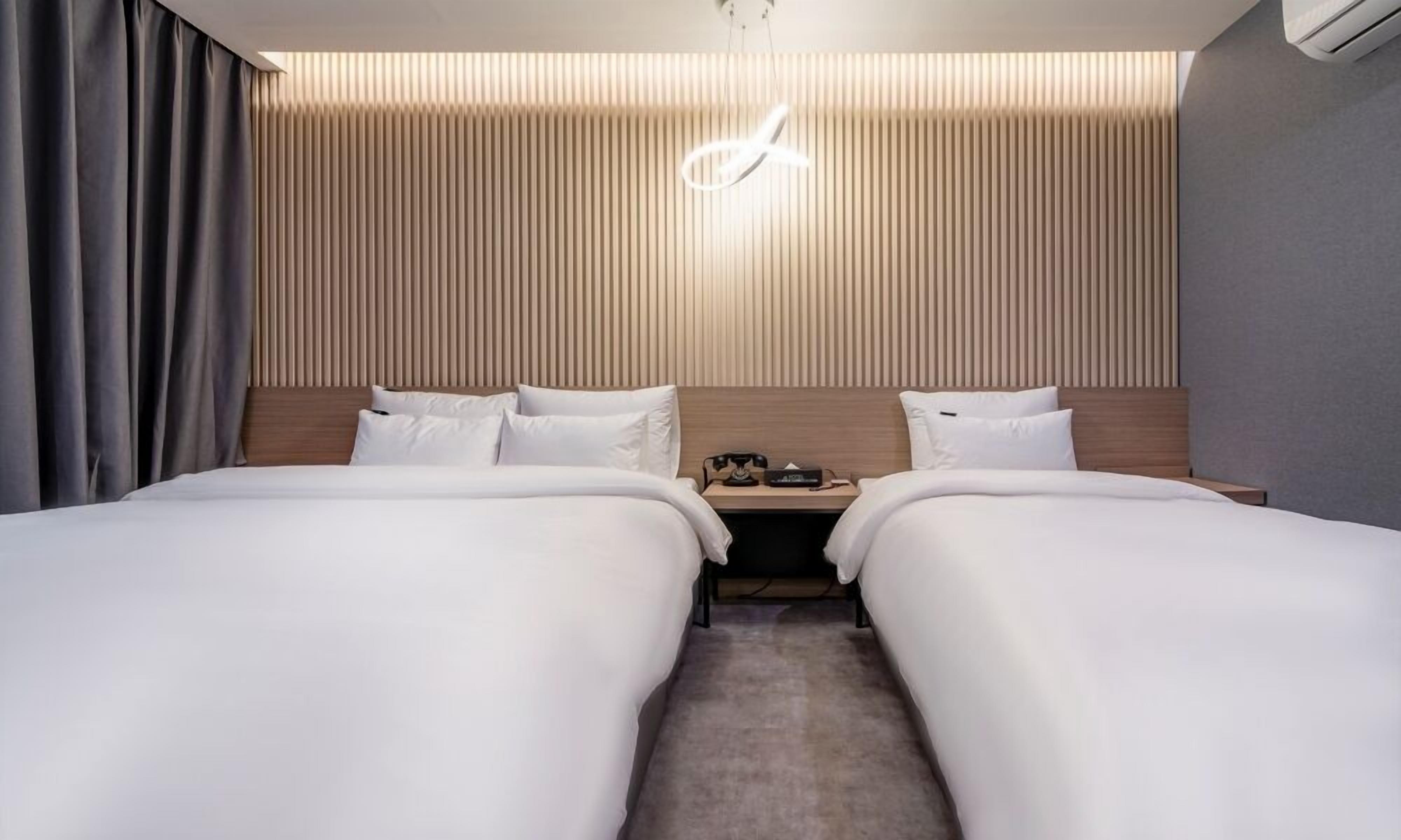 Quarto de hotel com duas camas, cabeceira de madeira, mesa de cabeceira com telefone e luminária moderna.