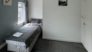 Een slaapkamer met een bed, een raam met gordijnen, een deur en een klein schilderij aan de muur.