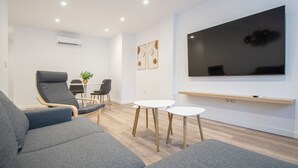 Uma sala de estar moderna com sofás cinzas, uma TV de tela plana fixada na parede e um pequeno espaço de jantar com mesa e cadeiras.