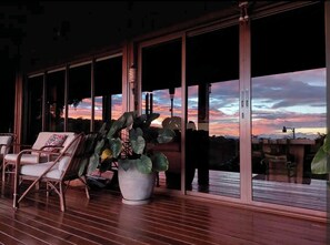 A spacious balcony with a view of a sunset over a mountainous landscape. There are wicker chairs and a large potted plant.