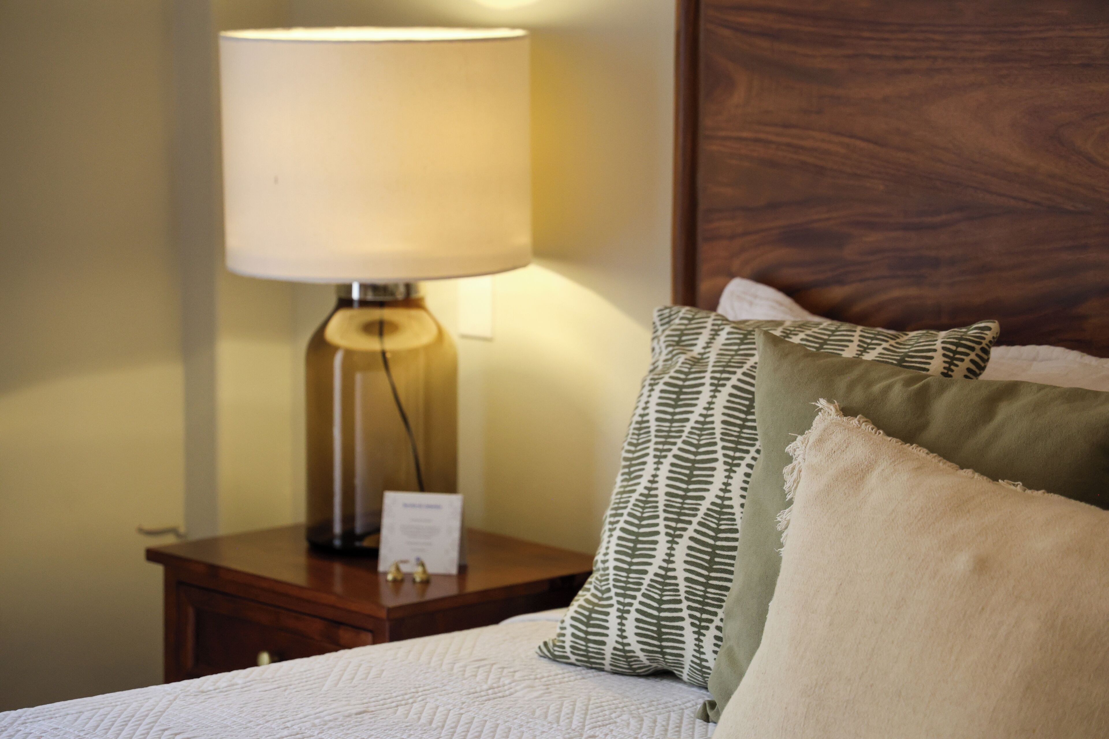 Een netjes opgemaakt bed met een witte quilt, een houten nachtkastje met een lamp en twee gestreepte kussens.