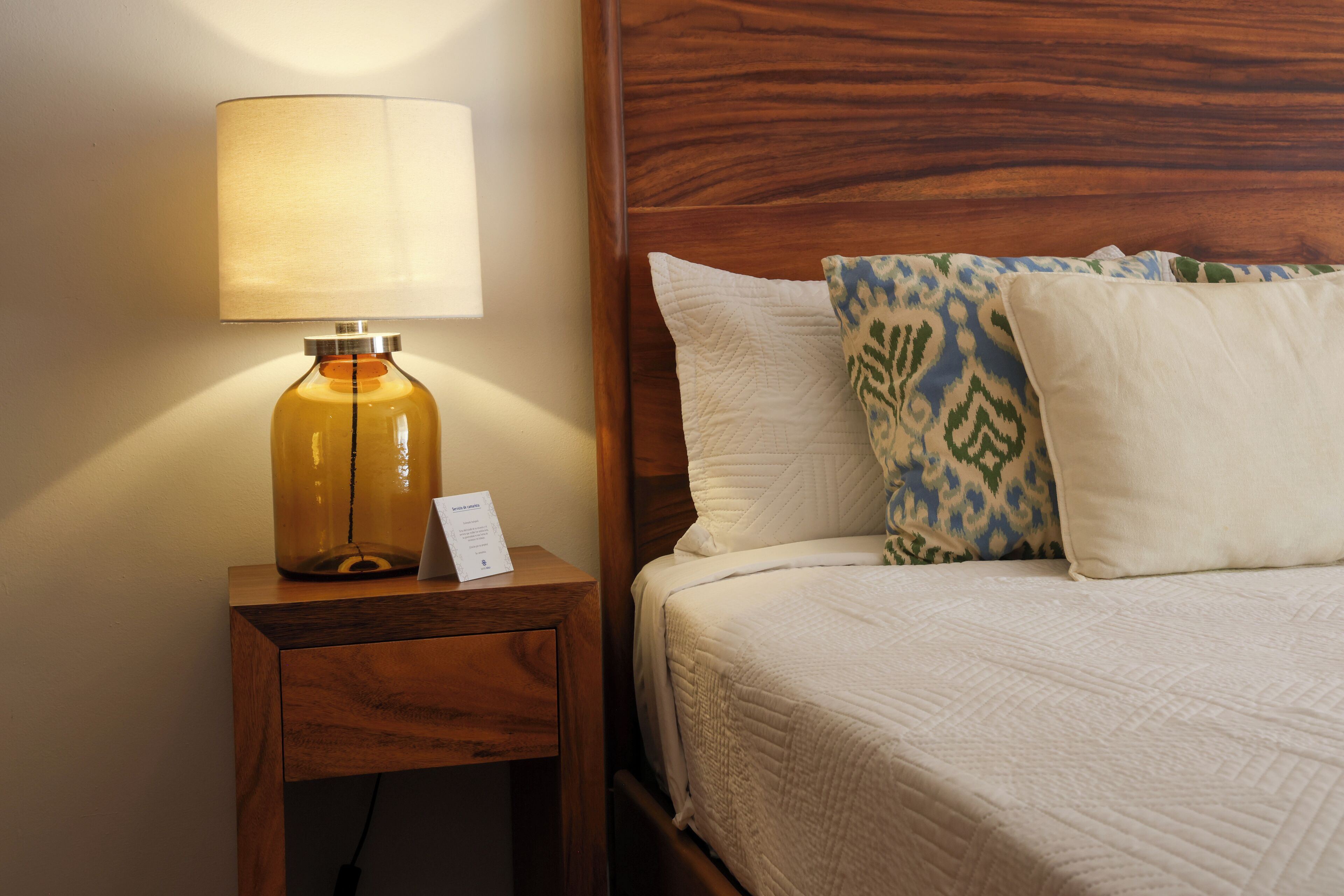 A neatly made bed with a white quilt and patterned pillows, a wooden nightstand with a lamp, and a wooden headboard.
