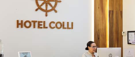 Een receptiebalie bij HOTEL COLLI, met een houten scheepsschijf logo, een bordje dat WiFi aangeeft en een ingelijst document aan de muur.