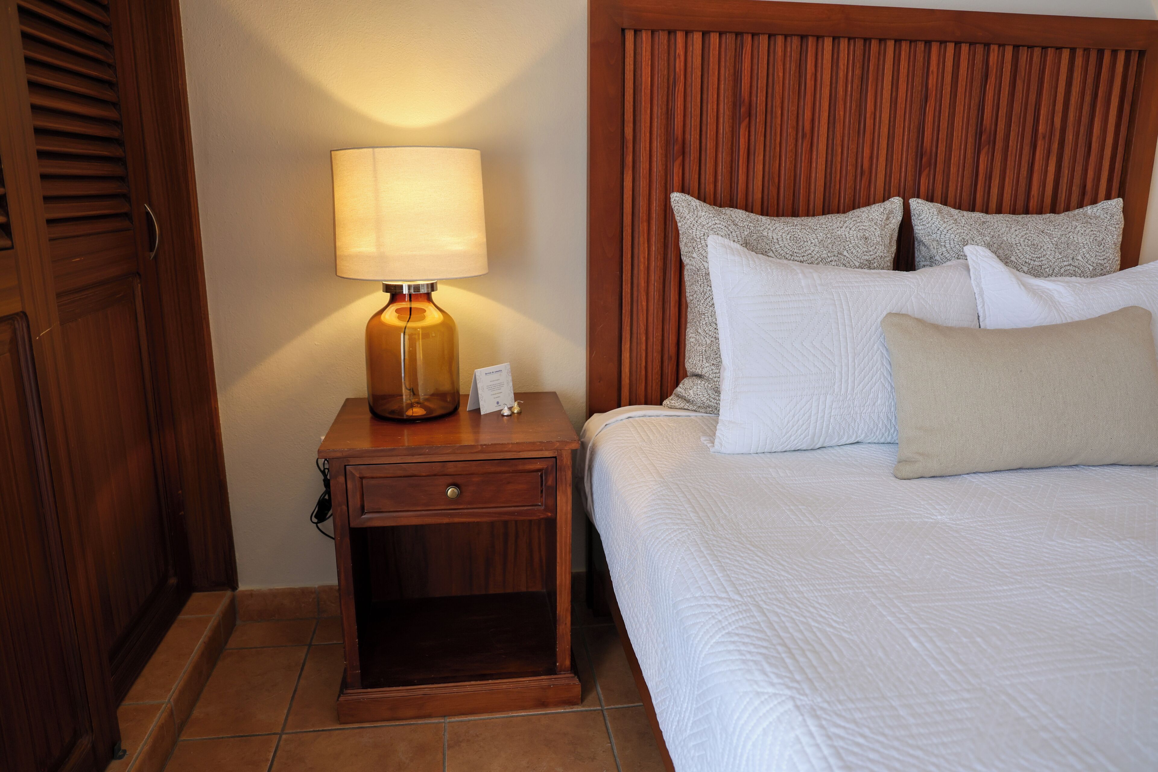 A neatly made bed with white linens and multiple pillows, a wooden nightstand with a lamp, and a wooden headboard.