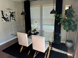 Moderni ruokailutila, jossa on lasipöytä, kaksi pehmustettua tuolia, mustat verhot ja ruukkukasvi.