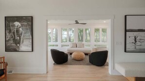 A modern living room with a sofa, two black chairs, a round woven ottoman, and a large window with a view of greenery. - Luxury Waterfront Cottage with Panoramic Waterway Views (Wilmington)