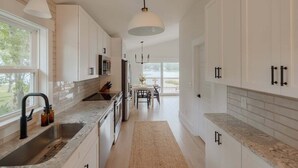 A modern kitchen with white cabinets, granite countertops, a large window, and a dining area with a view. - Luxury Waterfront Cottage with Panoramic Waterway Views (Wilmington)