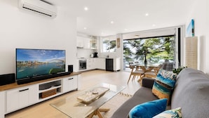 A modern living room with a flat-screen TV, a glass coffee table, and a grey sofa with colorful cushions.