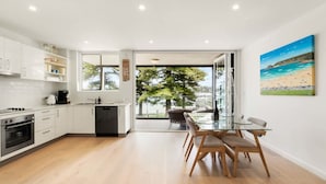 A modern kitchen with white cabinets, a dining area with a glass table and wooden chairs, and a large window with a view of a beach.
