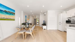 A modern kitchen with white cabinets, a dining area with a glass table and wooden chairs, and a beach-themed painting on the wall.