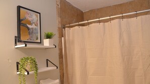 Un baño con cortina de ducha color beige, una obra de arte enmarcada sobre una repisa, y una planta en maceta en una estantería.
