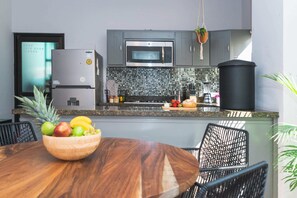 A modern kitchen with a microwave, refrigerator, and a black trash bin. There is a wooden bowl with fruits on a table in the foreground. - Casa Alonso | Spacious House with Private Courtyard in San Jose (San José del Cabo)