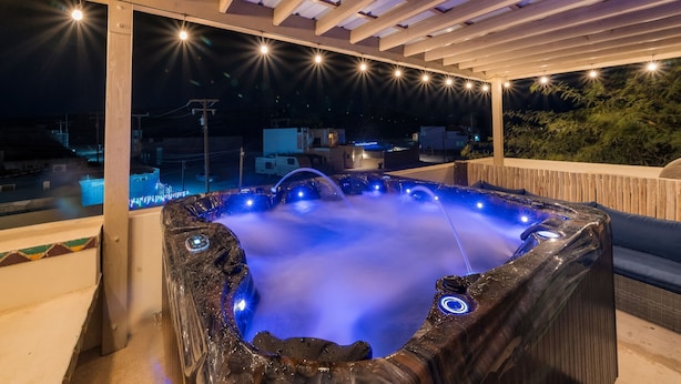 A hot tub under a pergola with string lights, surrounded by a wooden deck and overlooking a residential area. - Beach Home | Steps to Sand | Pool & Jacuzzi Unit A (Bahía la Choya)