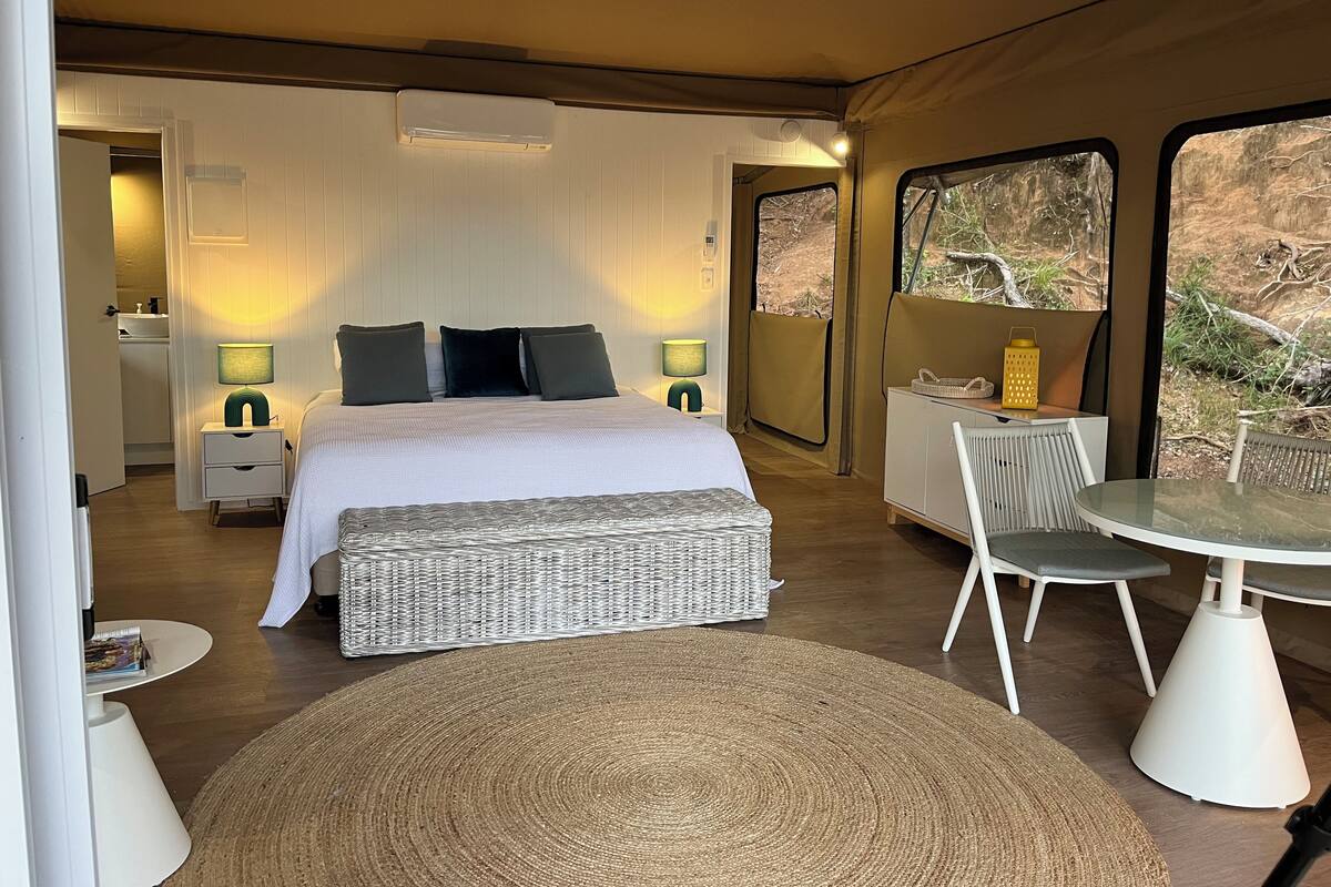 Ein Schlafzimmer mit Bett, Nachttischlampen, einem kleinen Tisch und einem Sessel. Ein Fenster bietet einen Blick ins Freie.