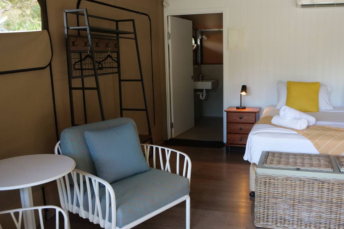 Ein Hotelzimmer mit Bett, Stuhl, kleinem Tisch, einem Korbwägelchen und einem durch eine offene Tür einsehbaren Badezimmer.