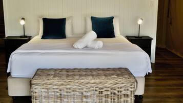A neatly made bed with white linens, two pillows, and a rolled towel on top. There is a wicker bench in front of the bed.