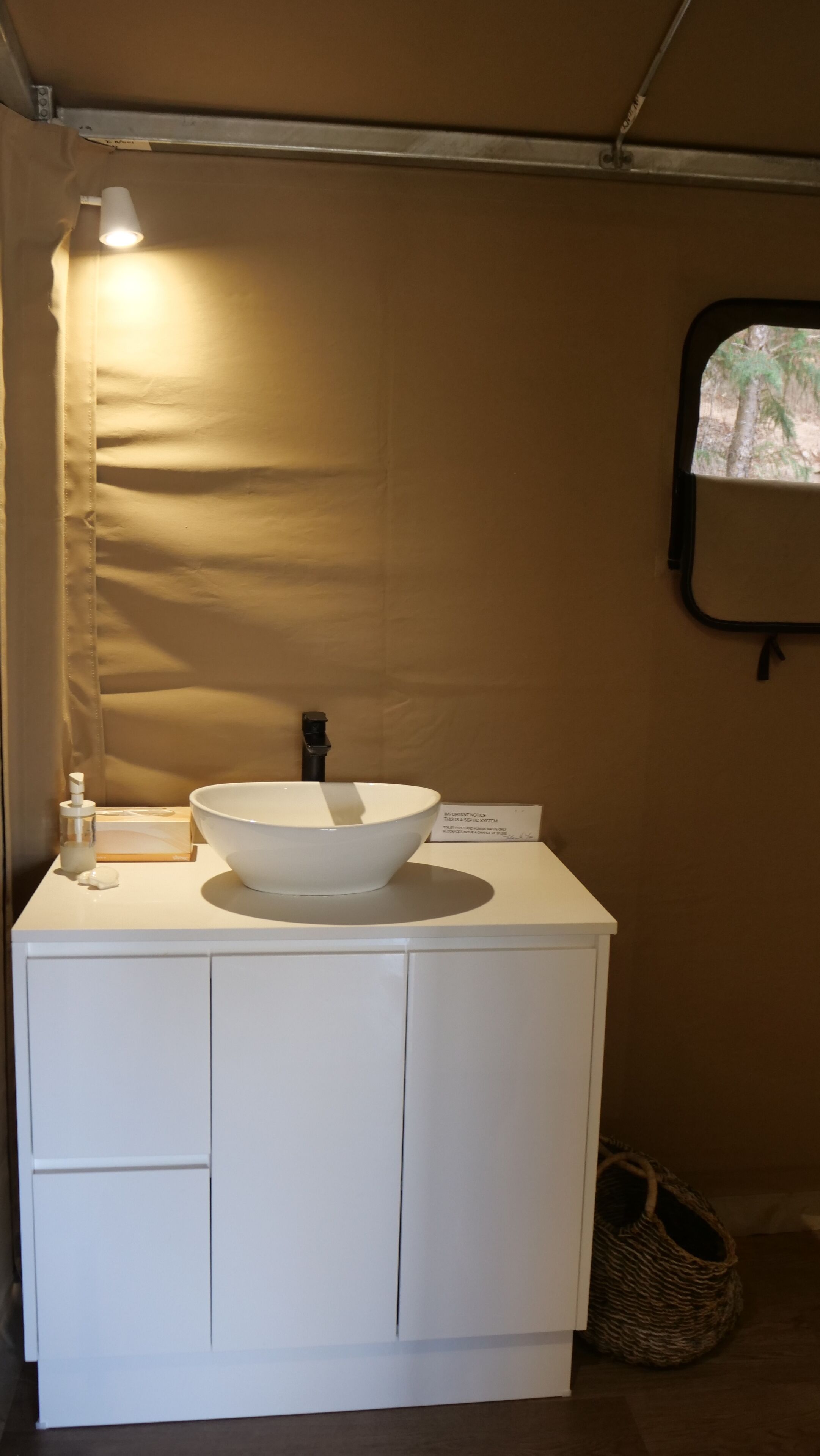 A white bathroom vanity with a large bowl sink, a handheld showerhead, and a small bottle of soap. There is a sign on the vanity and a window with a view of trees.