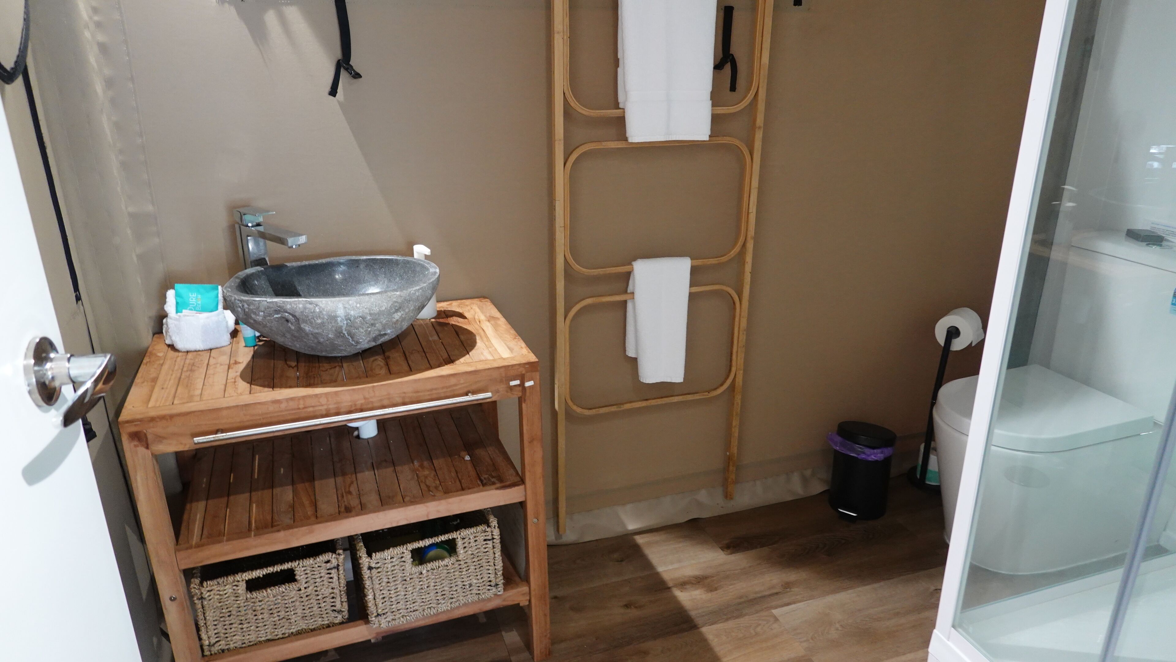 Een badkamer met een stenen wastafel, een houten wastafelmeubel, rieten manden, een handdoekhouder en een douchecabine met glazen wanden.