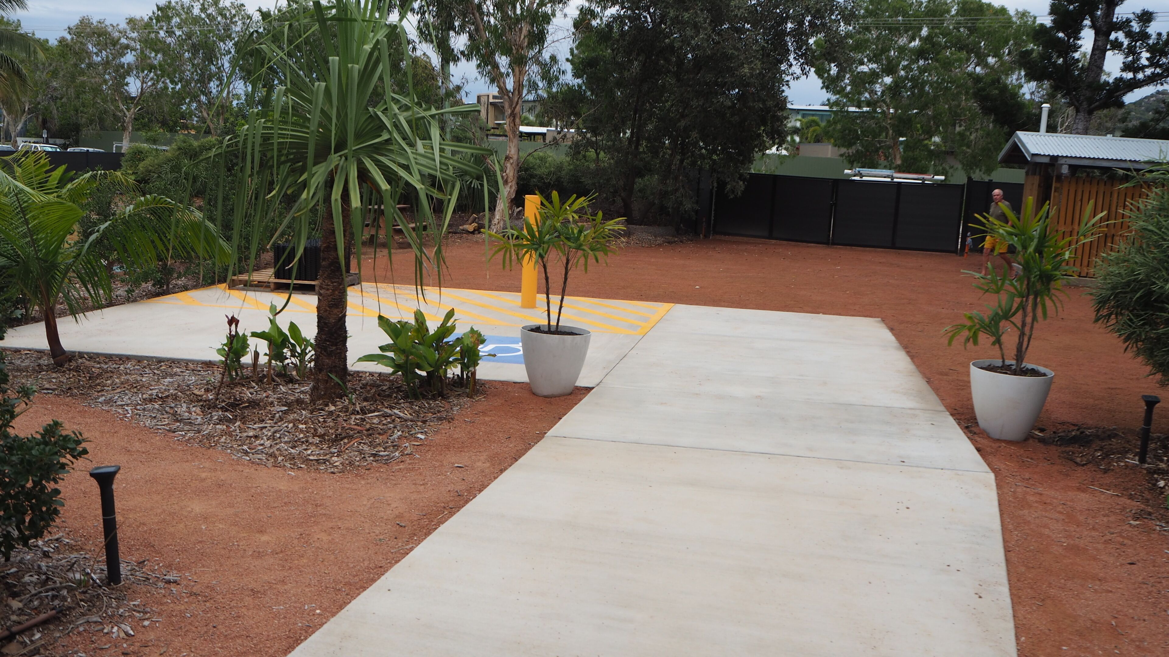 Un sendero pavimentado que conduce a un edificio con un área cercada que contiene diversas plantas y una superficie táctil amarilla.