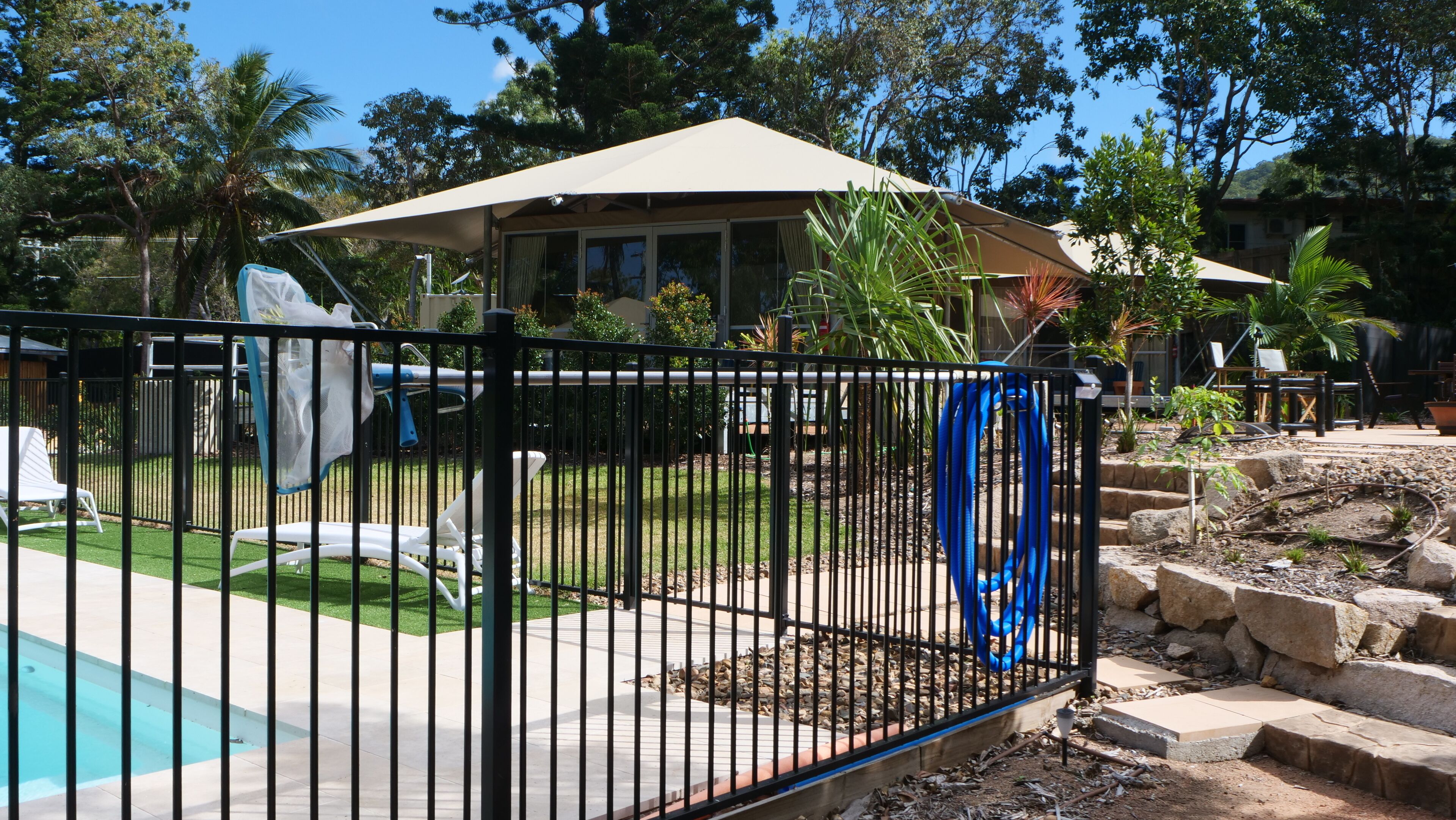 Un patio trasero cercado con piscina, un porche cubierto y una casa al fondo.