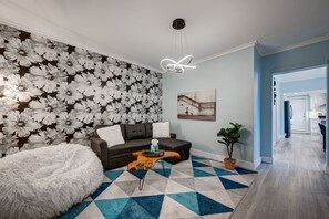 A living room with a floral wallpaper, a dark sofa, a white fluffy chair, a geometric patterned rug, and a modern light fixture.