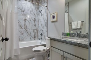 A bathroom with a marble shower wall, a bathtub, a toilet, a sink with toiletries, and a framed picture on the wall.