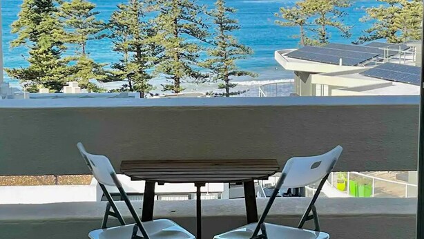 A balcony with a table and chairs, overlooking a coastal view with trees and the sea. - Beachfront Living, Manly Beach, Sydney (Manly)