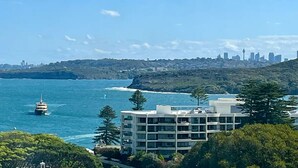 A coastal cityscape with a ferry in the water, a modern building with multiple floors, and a distant city skyline. - Beachfront Living, Manly Beach, Sydney (Manly)