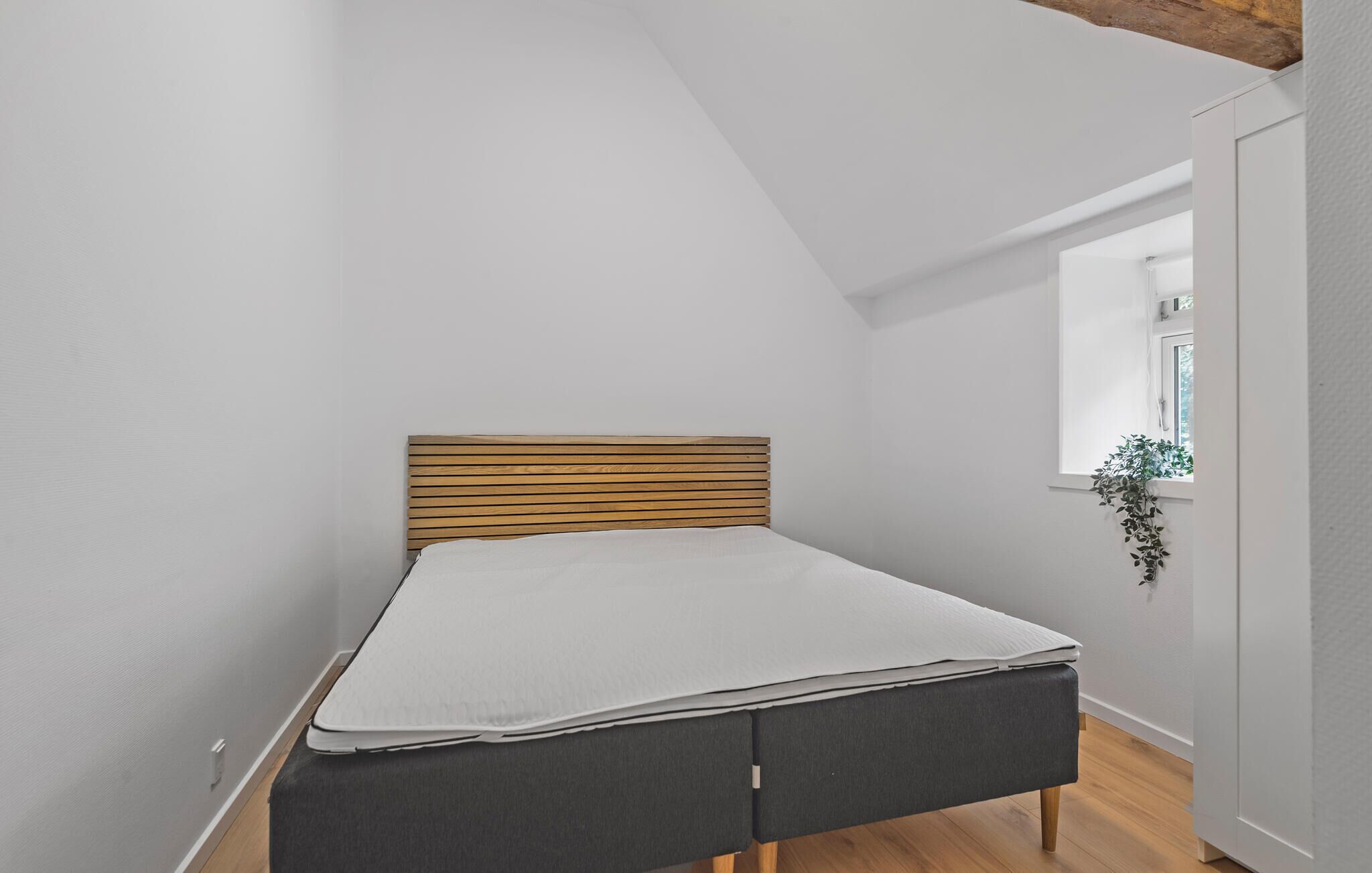 Une petite chambre à coucher moderne, dotée d’une tête de lit en bois à lattes, d’un matelas gris et d’un banc recouvert d’un tissu gris.