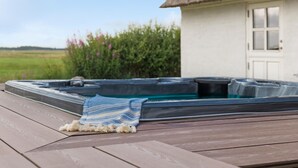 A hot tub on a wooden deck with a towel, a white building with a thatched roof, and a green field in the background. - Nice home in Janderup Vestj with sauna (Janderup Vestj)
