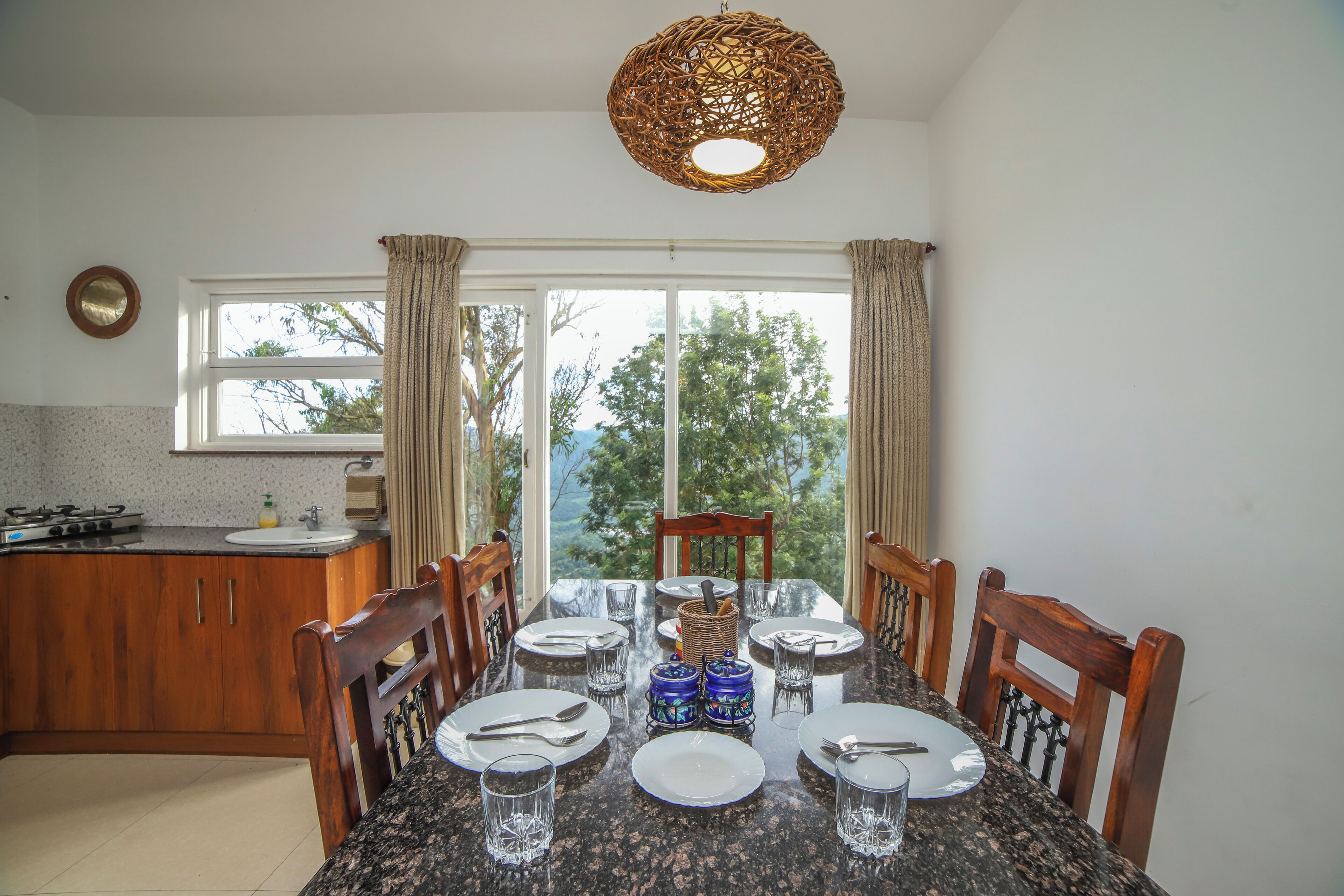 A dining area with a granite table set for four, wooden chairs, a view through large windows, and a kitchen in the background.