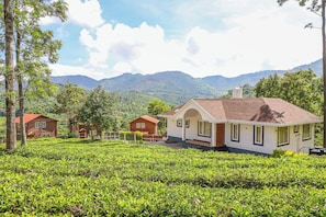 A house with a red roof and white walls, surrounded by lush green tea fields and mountains in the background. - Samrakshitha - The Villa (Kotagiri)